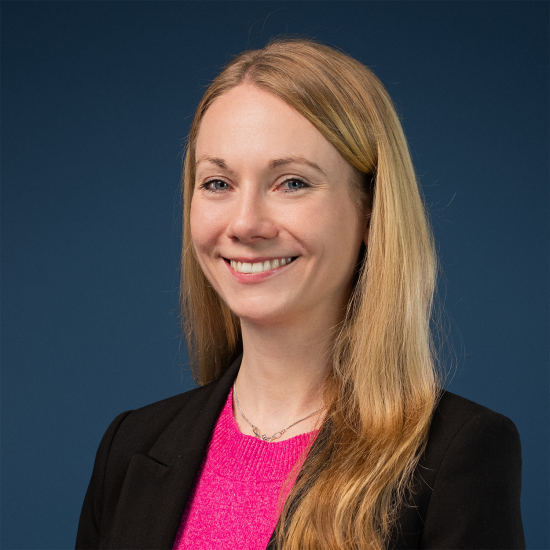 Porträt von Katharina Haen, die lächelnd vor einem blauen Hintergrund steht. Sie hat langes blondes Haar und trägt einen pinken Pullover sowie einen schwarzen Blazer.